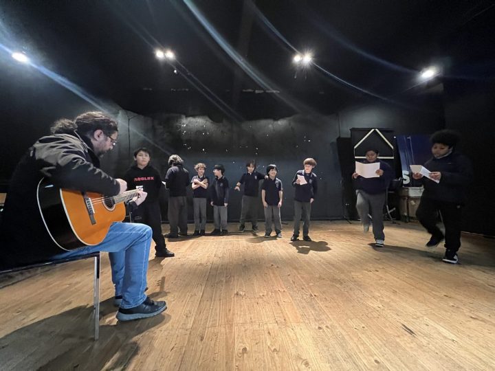 Estudiantes ensayando en el escenario con la letra en la mano, acompañados en guitarra